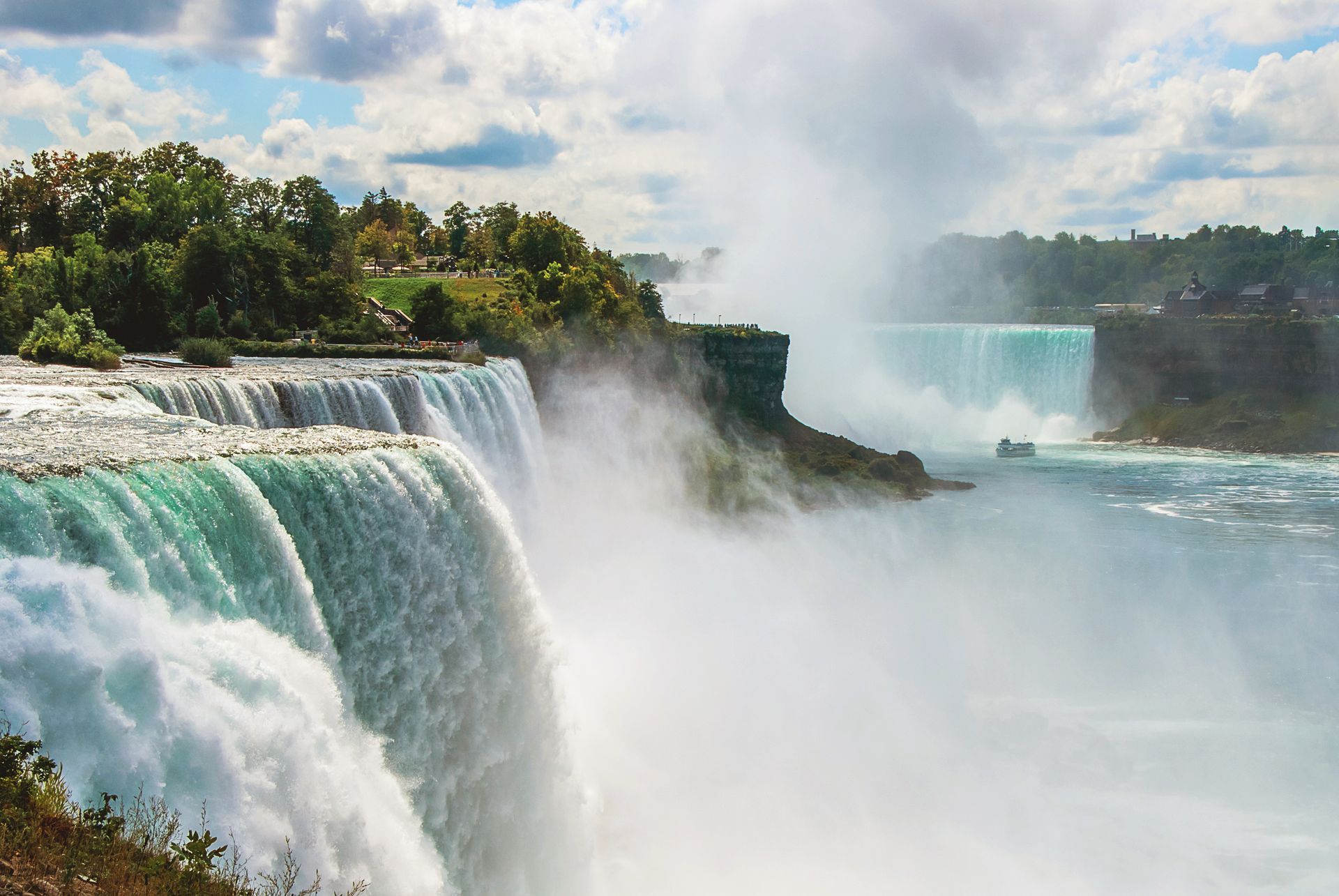 Massive waterfall with turquoise water and mist, viewed from a rocky, tree-lined cliff under a cloudy sky
