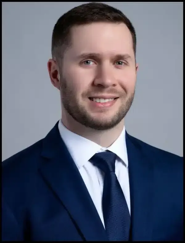 Studio headshot of a smiling person in a navy suit, white shirt, and tie against a gray background