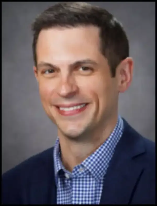 Studio headshot of a smiling man in a navy blazer and blue checkered shirt