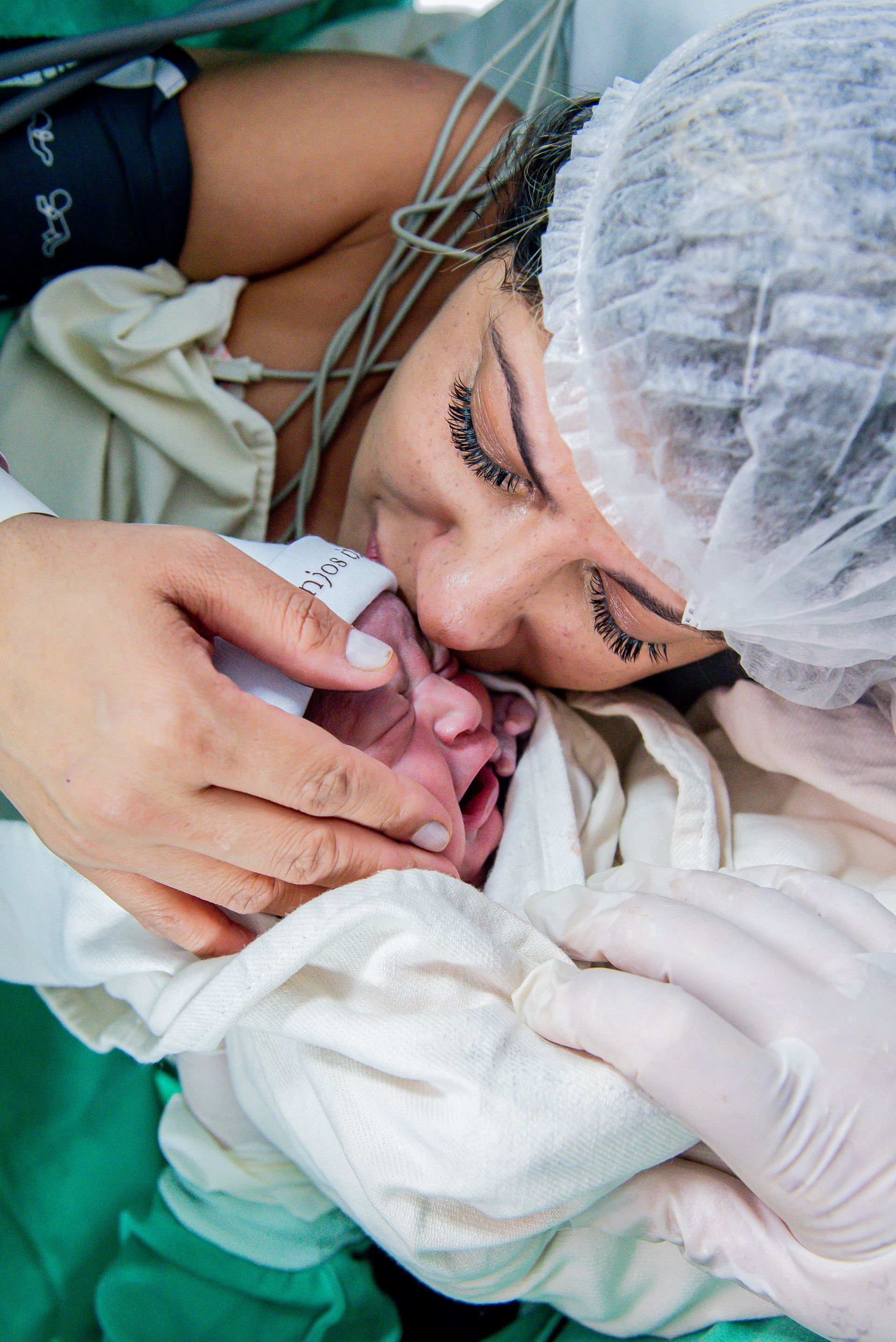 Newborn wrapped in blankets, held by gloved hands as a woman in a surgical cap kisses the baby’s forehead