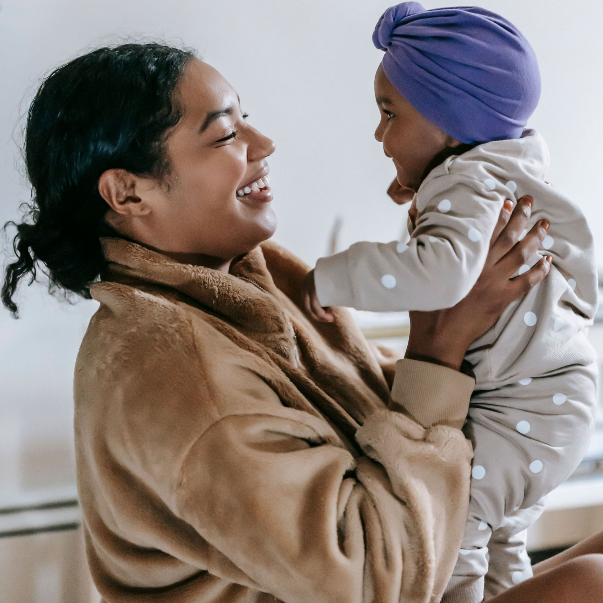 Woman smiling at a baby in a purple turban and polka-dot outfit, holding them in a cozy indoor setting