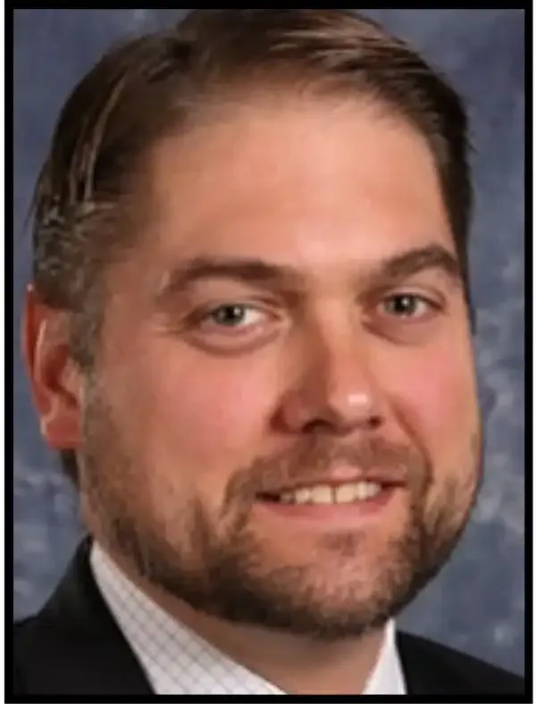 Close-up headshot of a man in a suit and tie, smiling against a dark background