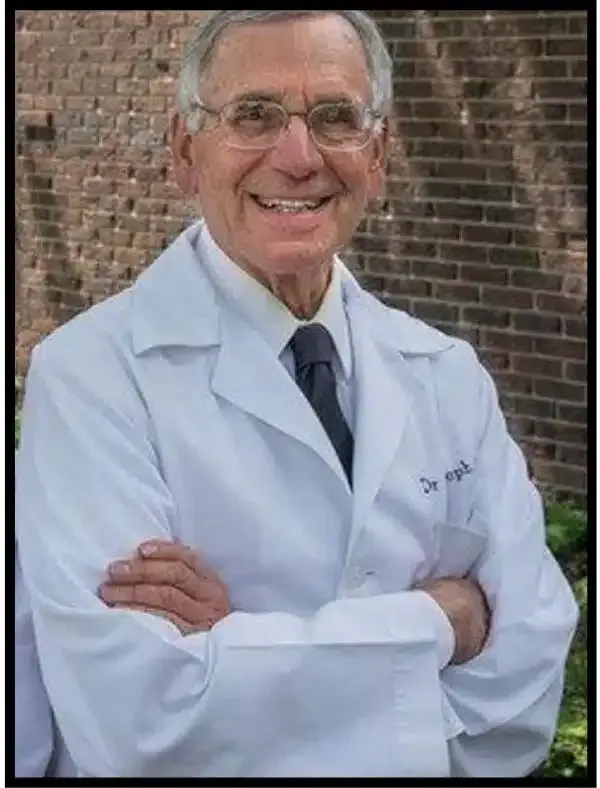 Smiling doctor in a white lab coat with arms crossed, standing in front of a brick wall.
