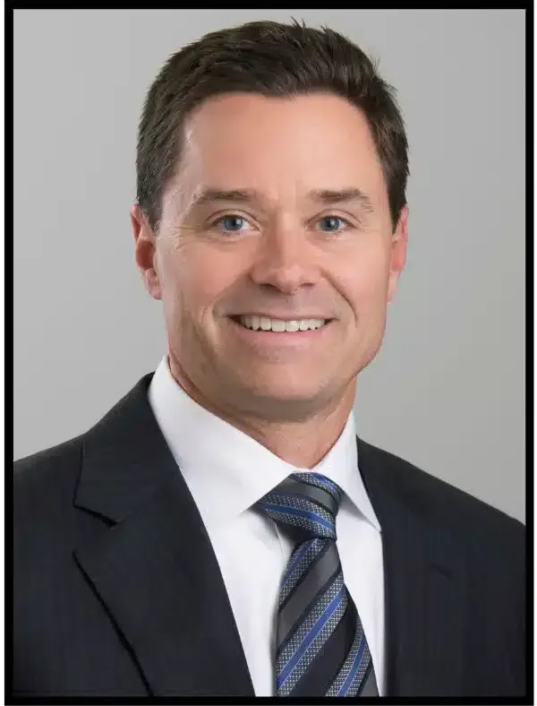 Professional headshot of a smiling person in a dark suit and striped tie against a gray background