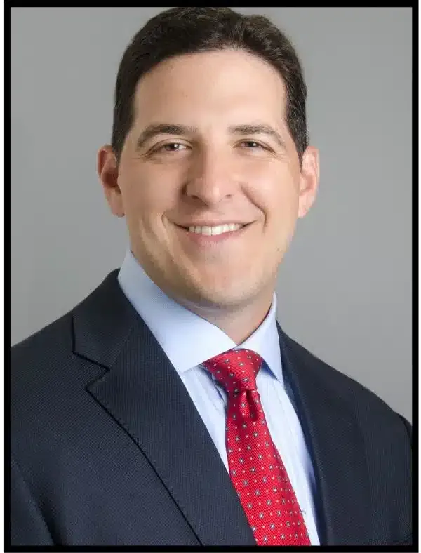 Studio headshot of a smiling person in a dark suit, light blue shirt, and red tie on a gray background