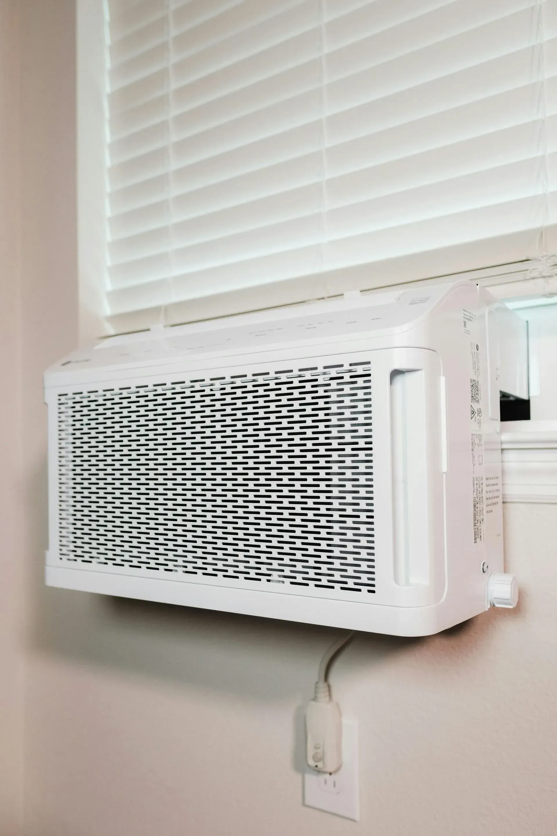 White window air conditioner unit installed in a window, with a power cord connected to a wall outlet.