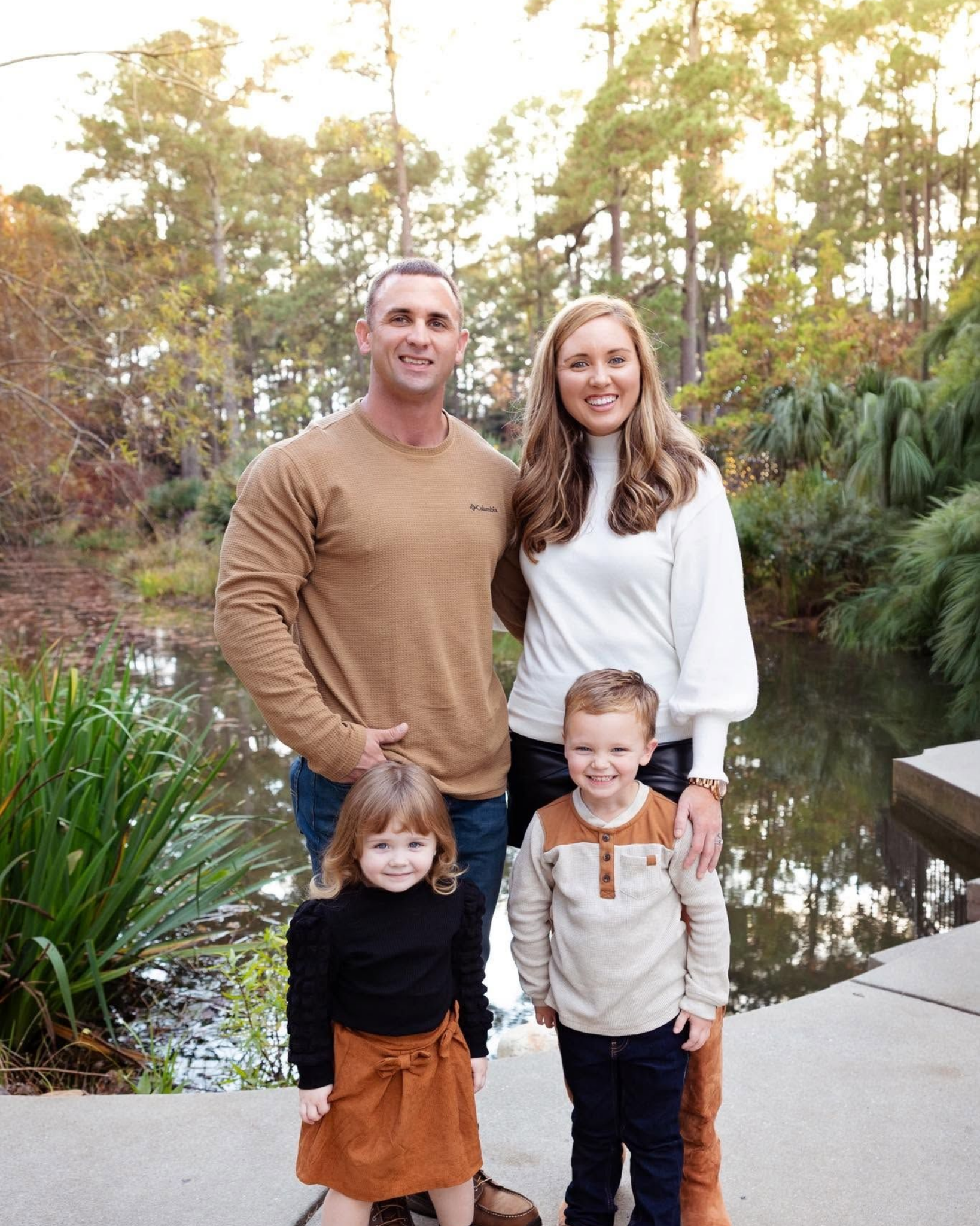 Family of four standing outdoors by a pond, with trees and greenery in the background.