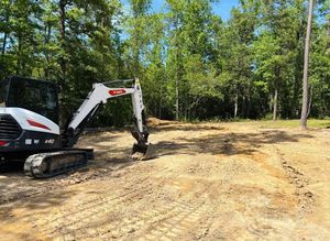 Excavator leveling a dirt lot beside a wooded area under a clear sky