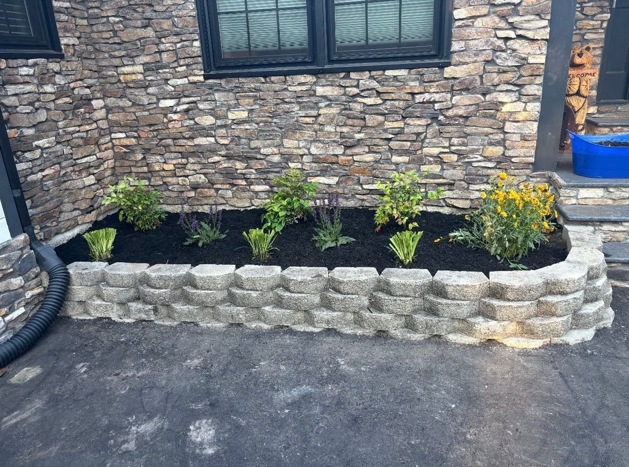 Stone planter bed with black mulch and small yellow flowers along a brick wall by a driveway