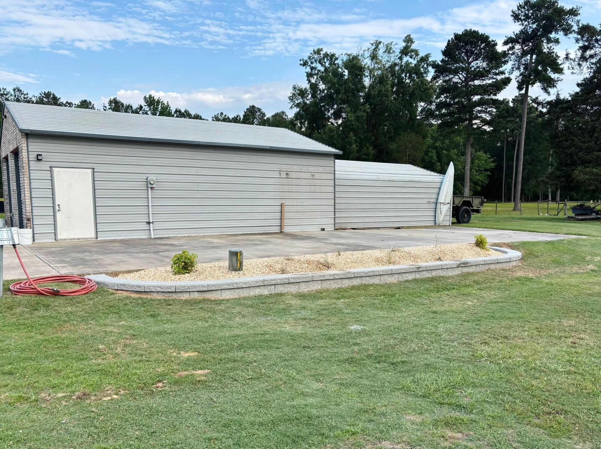 Gray metal building beside a gravel lot and grass, with trees in the background.