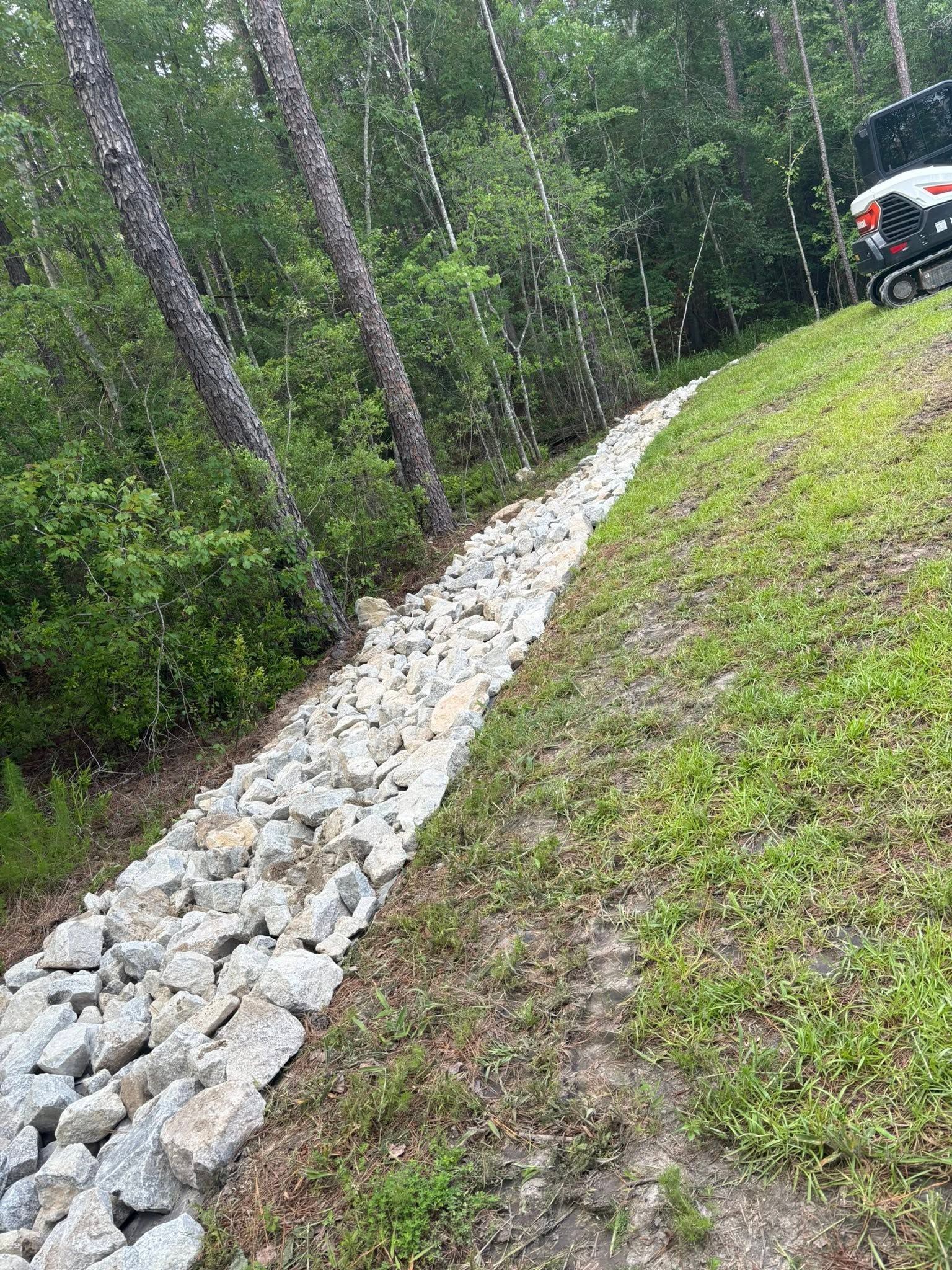 Rock-lined drainage swale through a grassy yard beside a wooded area, with an off-road vehicle at the edge.