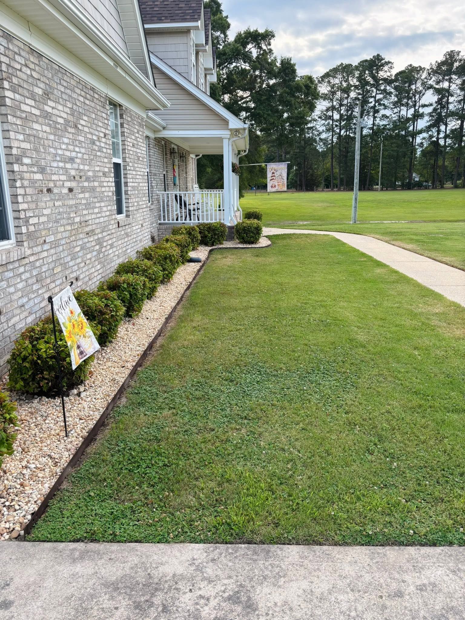 Side yard of a stone house with a green lawn, small shrubs, and a concrete walkway along the grass