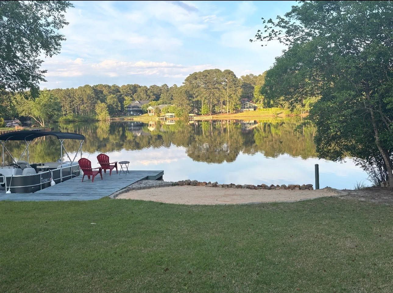 Lakeside dock with red chairs, calm water reflecting trees at sunset, framed by a grassy shore