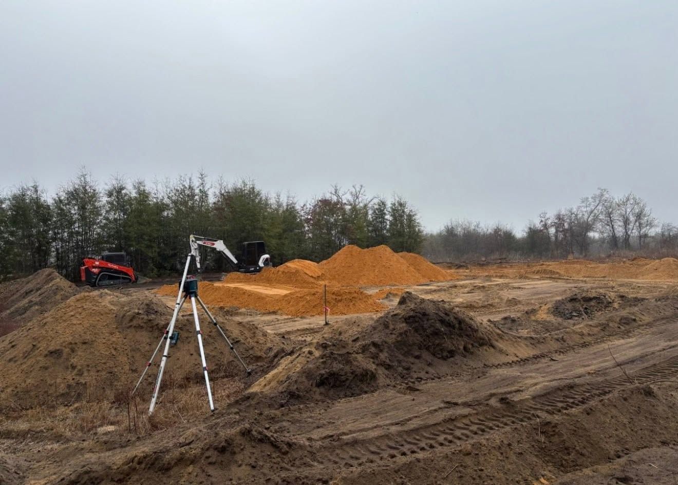 Construction site with orange dirt piles, a tripod survey marker, and an excavator under an overcast sky