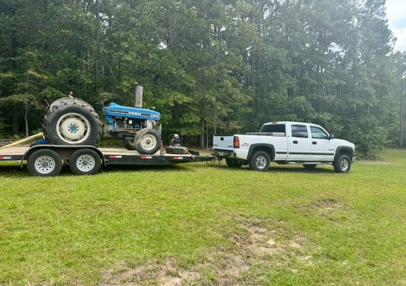White pickup towing a trailer with a blue tractor across a grassy field, with trees in the background