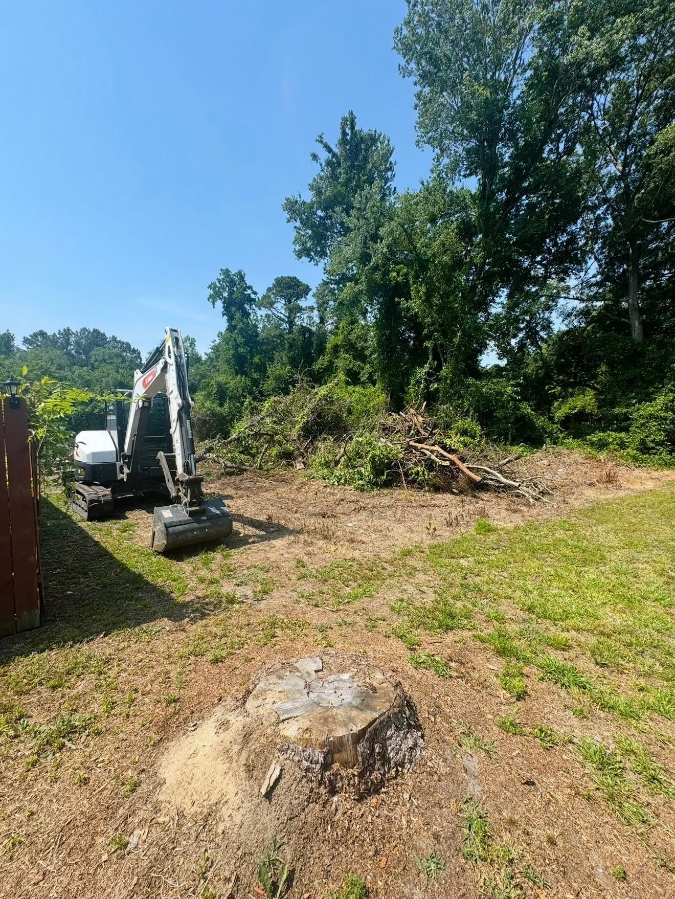 Excavator clearing brush on a sunny grassy lot with trees and piles of dirt.