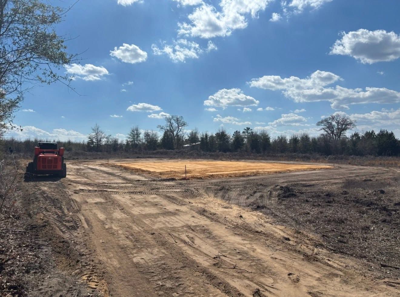 Dirt lot under blue sky with clouds, a red vehicle at left and trees along the horizon
