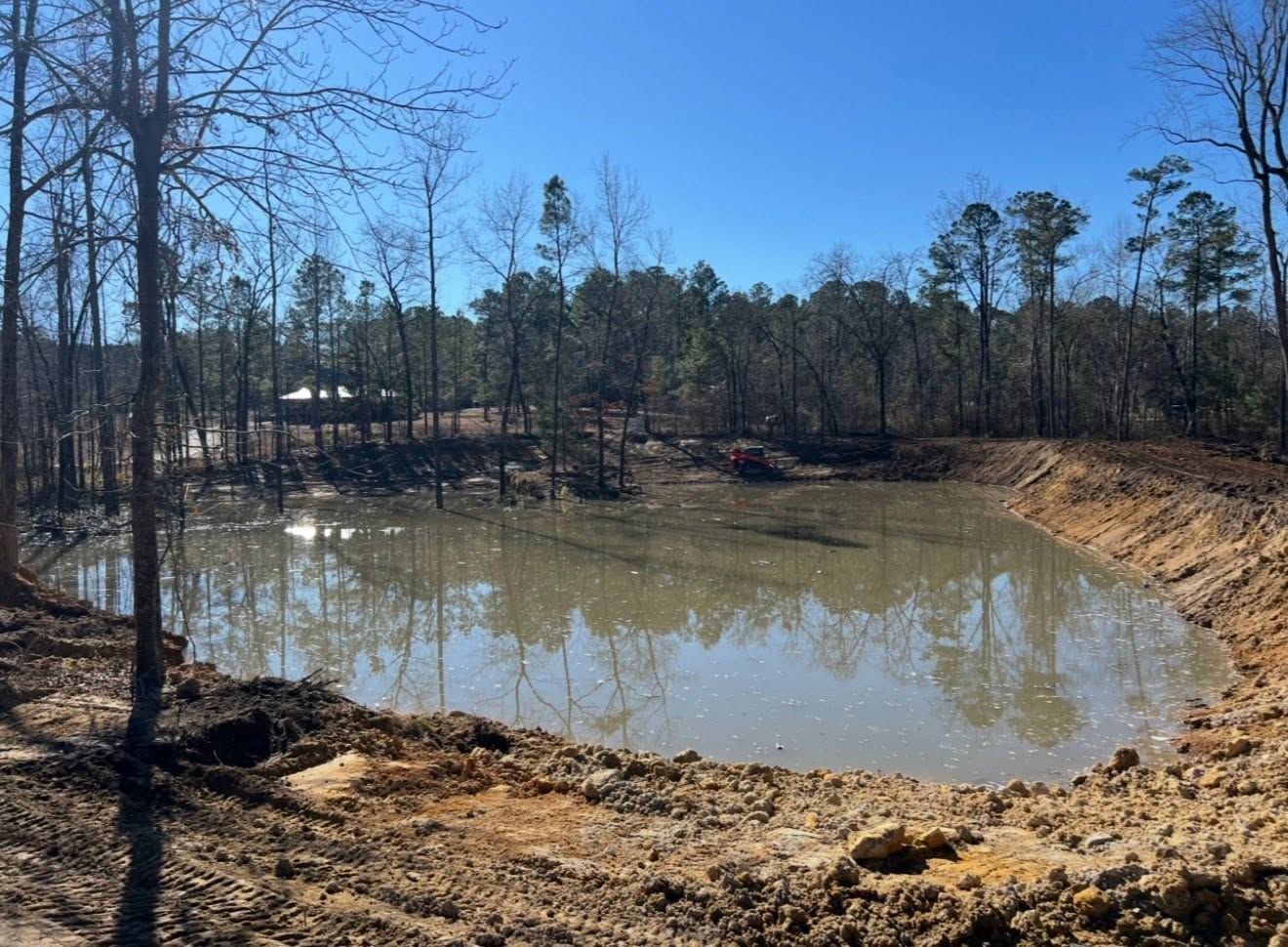 Small muddy pond or excavation basin with bare trees and wooded banks under a clear blue sky