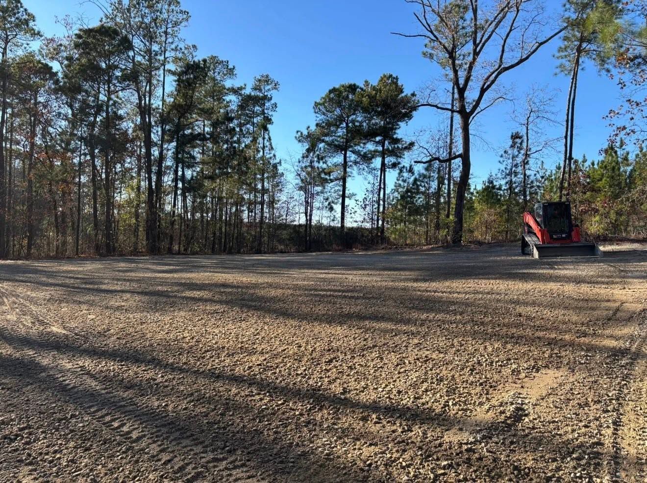Gravel lot beside a wooded area under a clear blue sky, with a red tractor at the right edge.