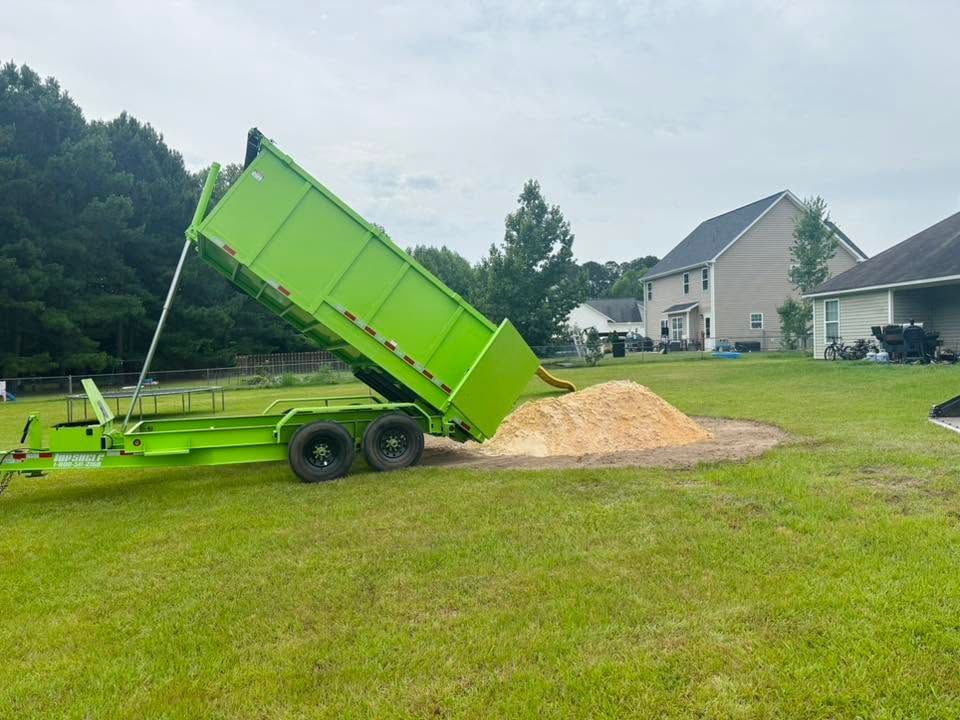 Bright green dump trailer tipped on a lawn, unloading a pile of mulch near suburban houses.