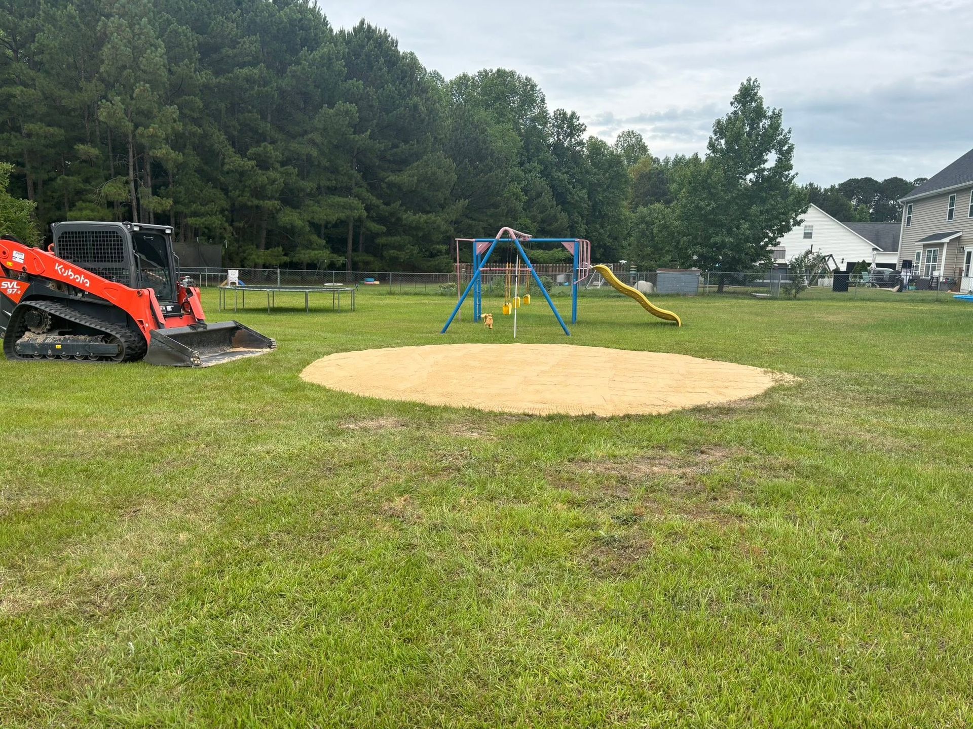 Empty playground with a blue swing set and yellow slide, beside an orange excavator on a grassy field.