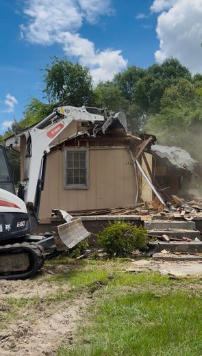 Partially collapsed house with debris and a parked vehicle under a bright blue sky