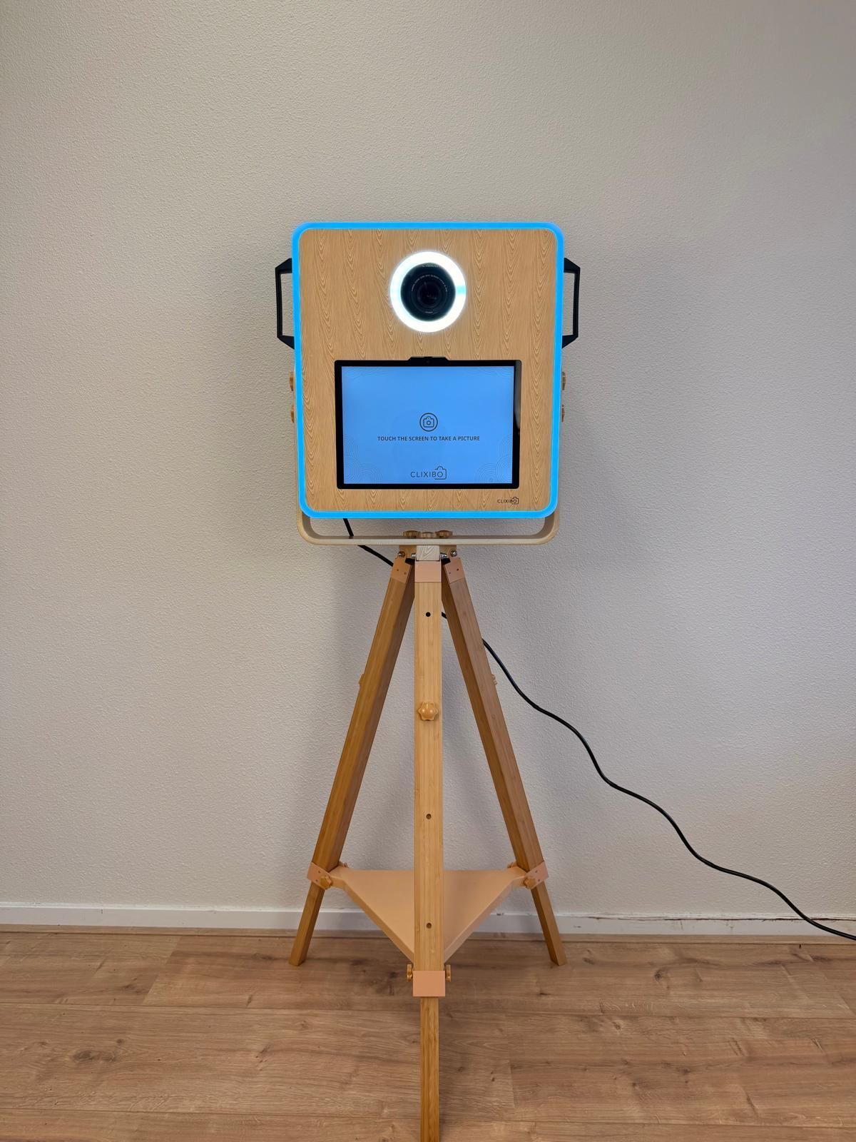 Een draagbare fotocabine met een tabletscherm en ringlamp, gemonteerd op een houten statief tegen een lichtwand. Photobooth