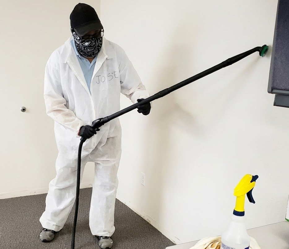 Worker in white protective suit using a long pole to clean a wall in a plain room.