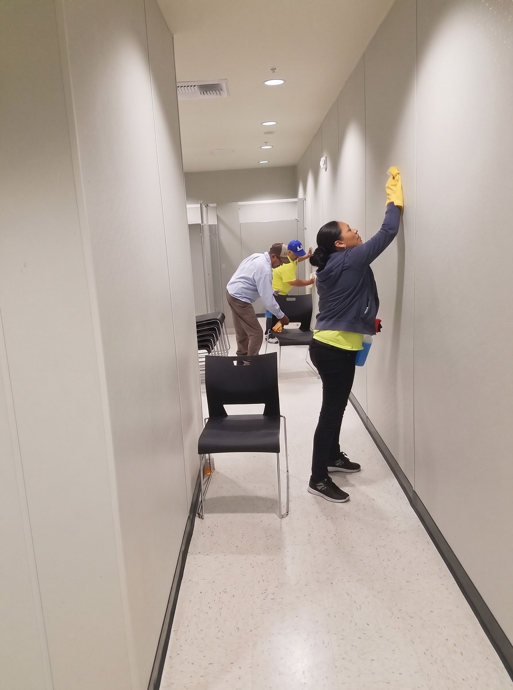 Two workers inspecting a narrow hallway, one reaching up to the wall, with tools and a chair nearby.