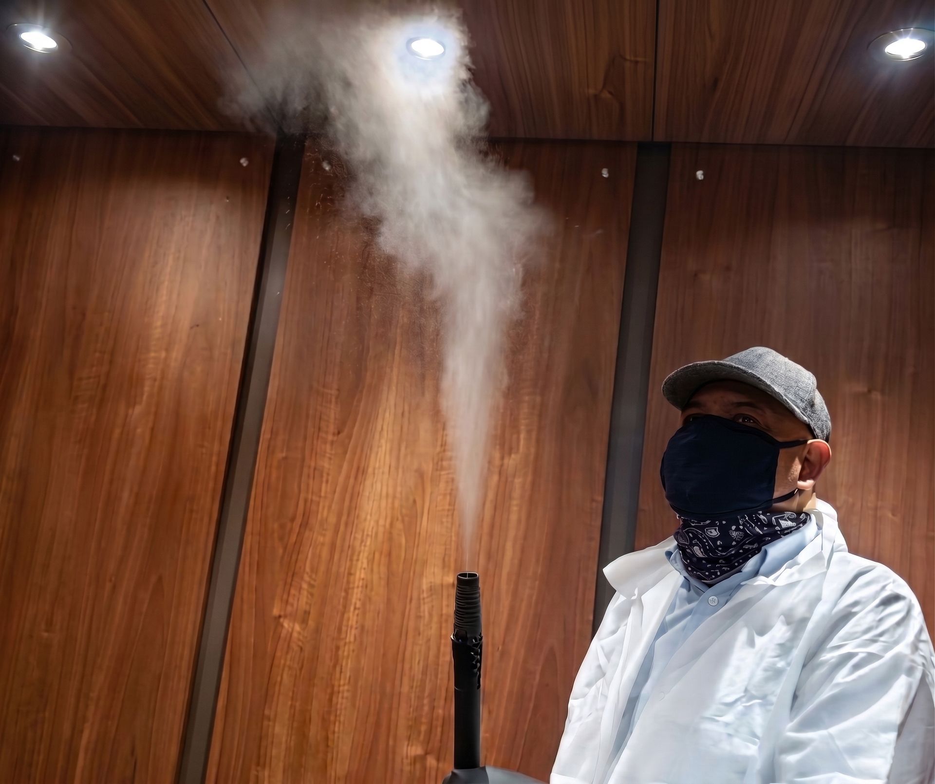Person in a white coat and cap operating a fog machine in a wood-paneled room.