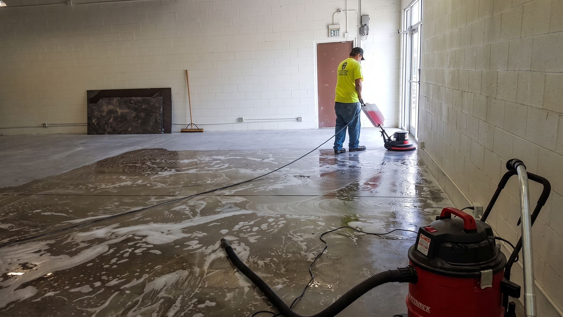 Worker cleaning a wet floor in a bare room with a red vacuum at right