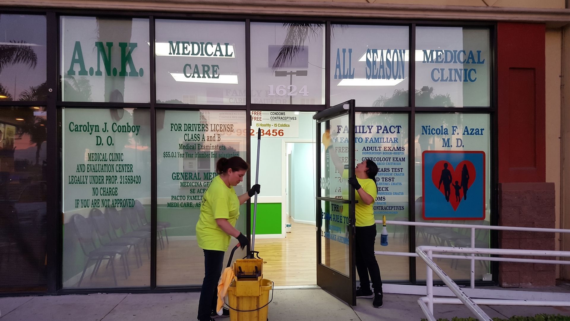 Two workers in neon safety jackets outside a medical clinic entrance, one using a yellow floor machine.