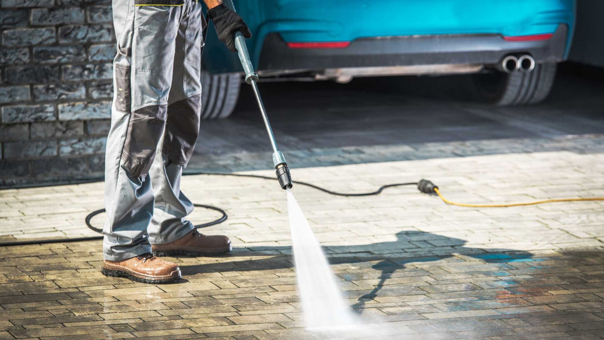 Person pressure-washing a driveway beside a blue car and stone wall.