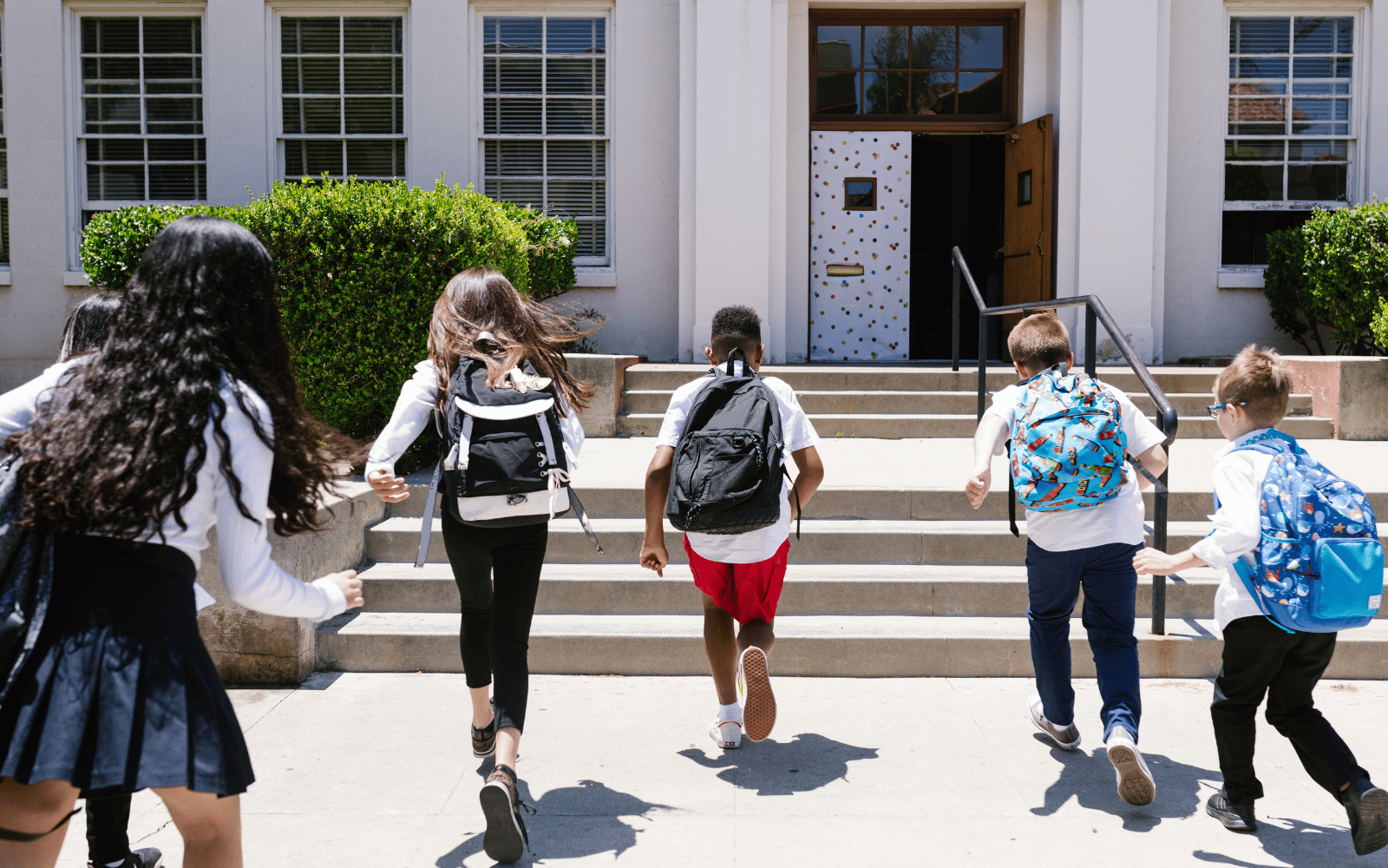 Students with backpacks running up the steps towards a school entrance.