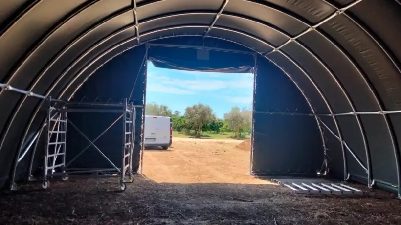 Vista interna di una grande struttura con tetto ad arco e porte aperte verso l'esterno. Un furgone parcheggiato in lontananza.