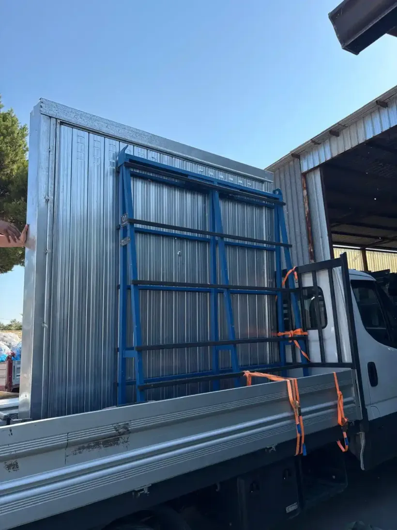 Un camion con pianale che trasporta una struttura in pannelli metallici con telaio blu, fissata con cinghie arancioni.