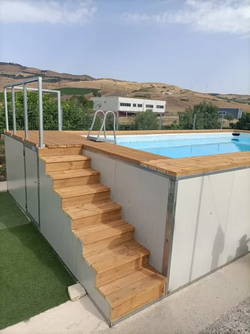 Piscina con gradini in legno, circondata da pareti e ringhiere dai colori chiari. Sullo sfondo una casa e delle colline.