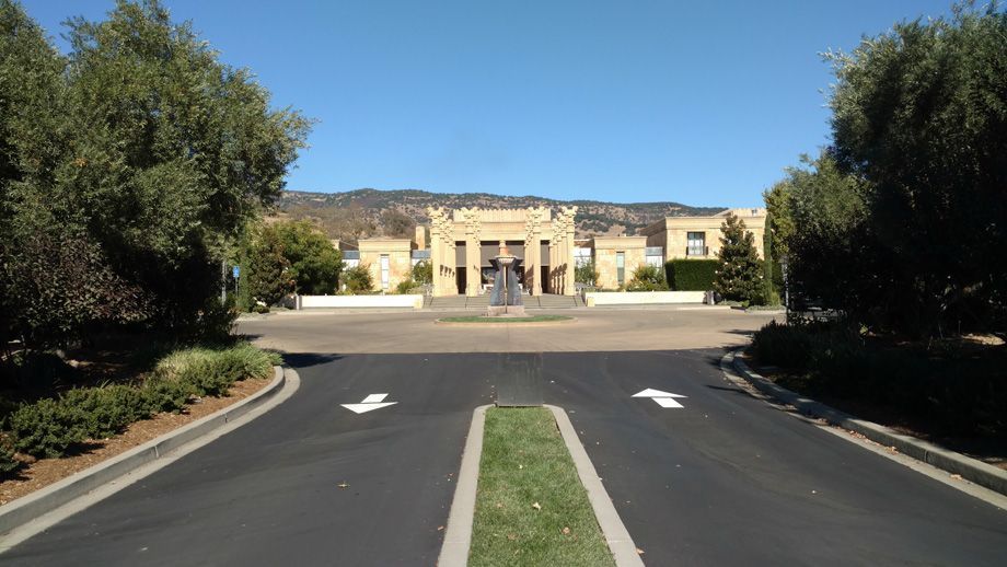 Driveway leading to a beige building with arched entrance, flanked by trees under a clear blue sky.