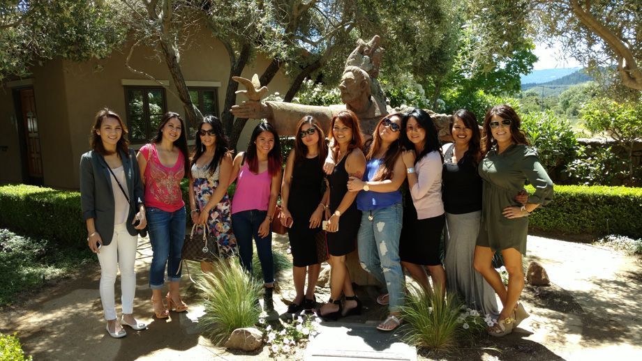 Group of people posing outdoors in front of a building, surrounded by trees and bright sunlight.