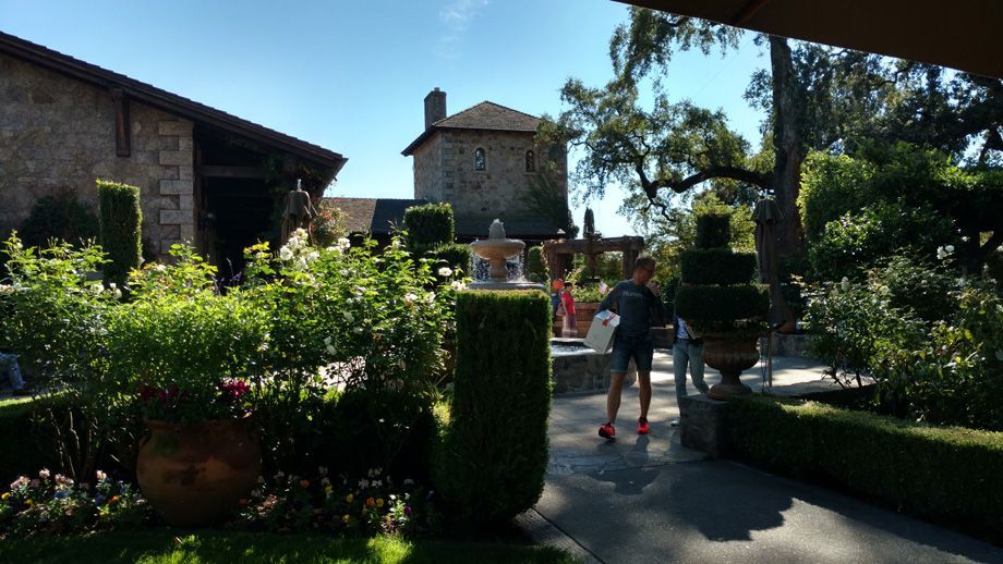 Sunny garden walkway with stone buildings, hedges, and two people near a path