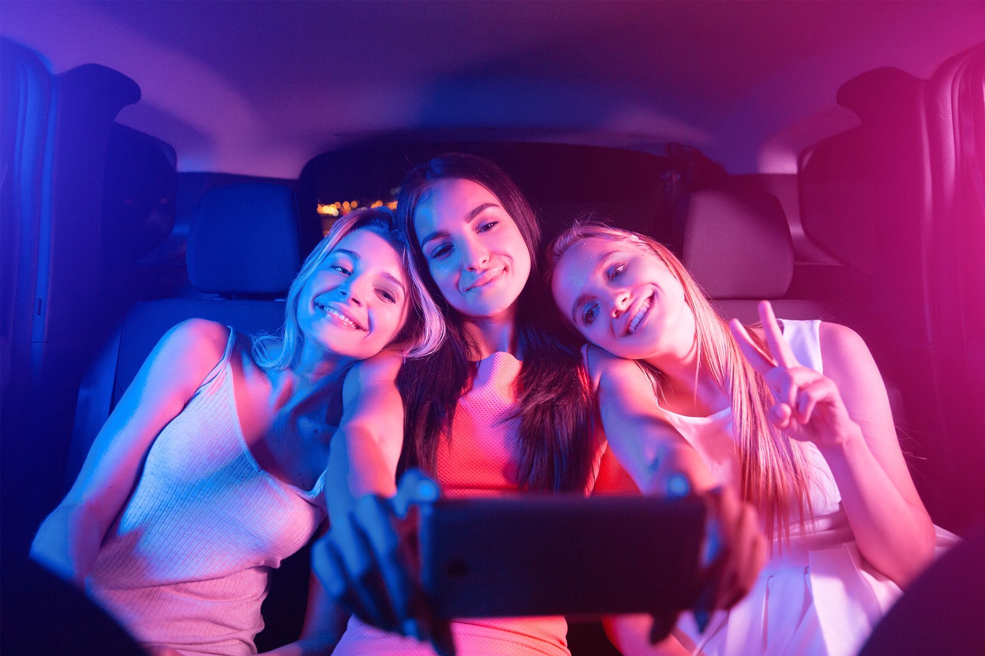 Three friends smiling in a car at night, lit by blue and pink neon light.