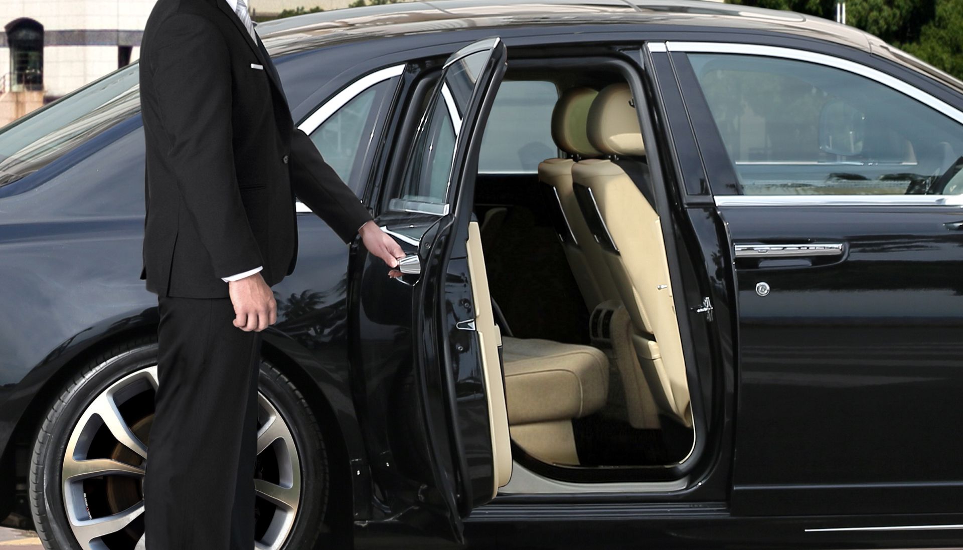Man in black suit opening the rear door of a black sedan, revealing beige interior.