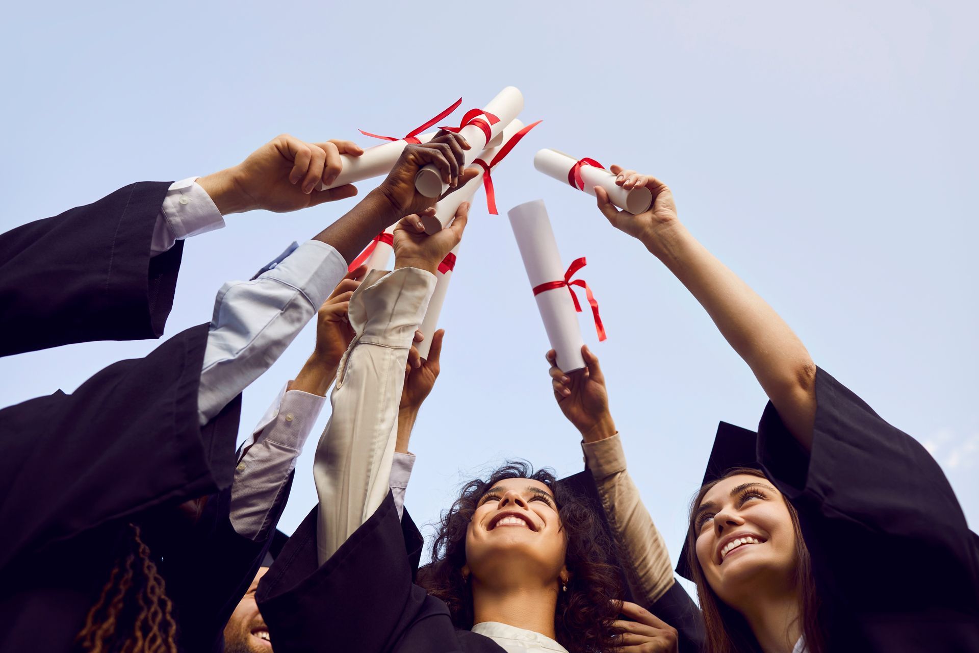 Graduates in caps and gowns raise diplomas overhead against a blue sky