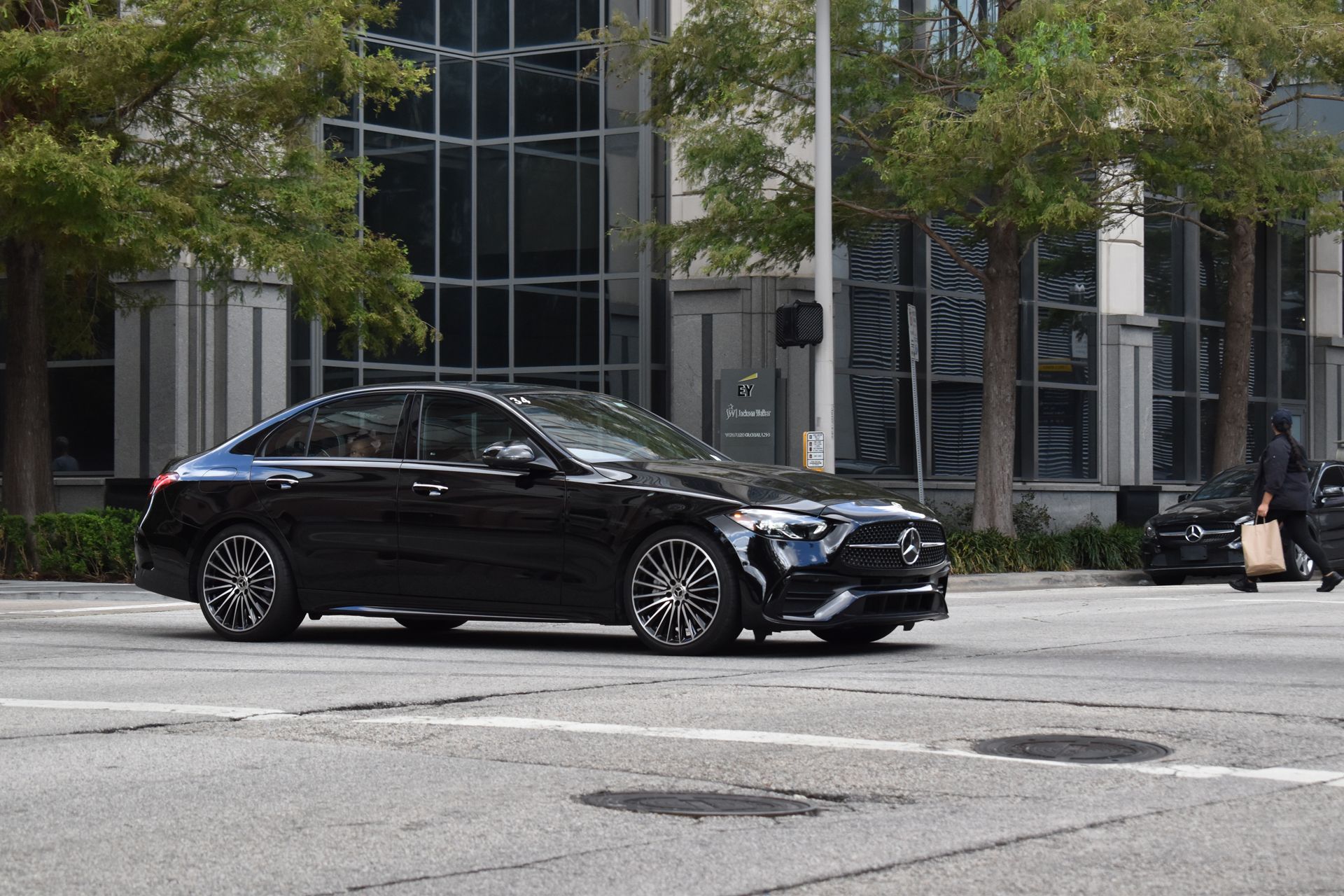 Black sedan parked on a city street in front of a glass office building, with trees lining the sidewalk.