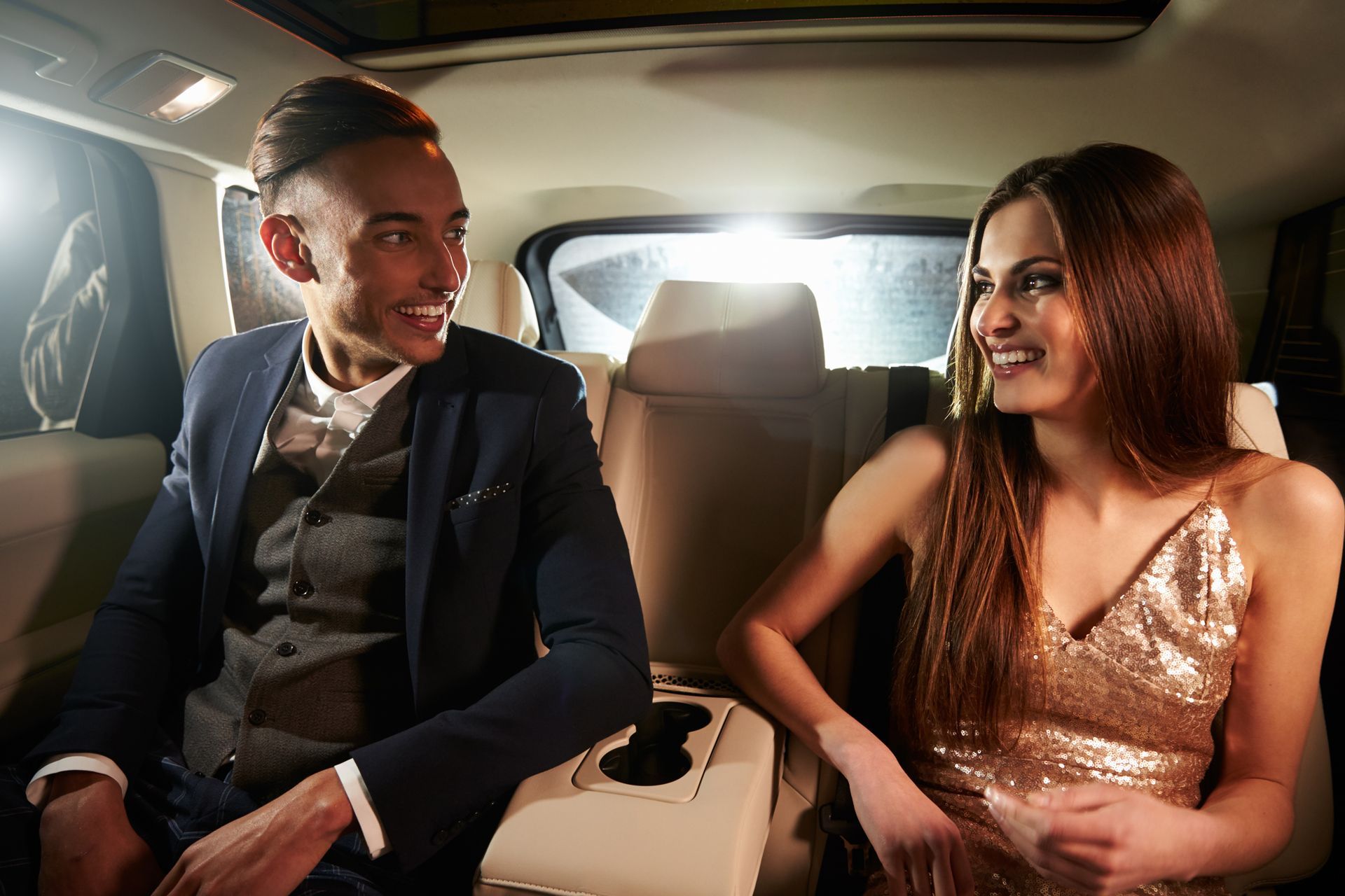 Two people in a luxury car smiling at each other, with a bright city light through the window.