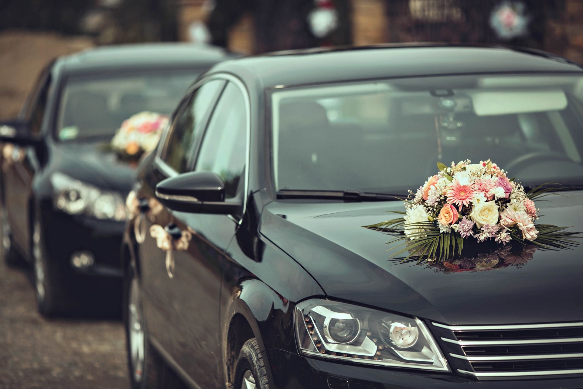 Black wedding car with floral bouquet on the hood, followed by another decorated car in a wedding procession