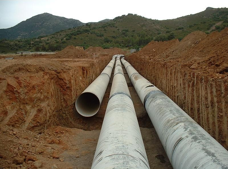 Tre grandi tubi posati in una trincea, preparati per la sepoltura in un ambiente di terra marrone con uno sfondo di montagne.