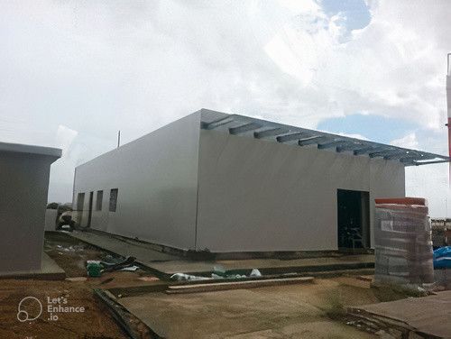 Edificio rettangolare bianco in costruzione, con travi metalliche lungo il bordo superiore e cielo coperto.