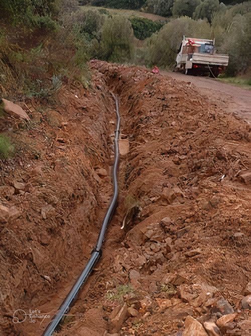 Trincea scavata nella terra con un tubo nero all'interno, camion sullo sfondo su strada sterrata, pendio della collina in lontananza.