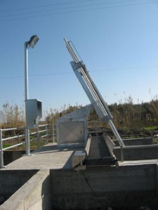 Sistema di trattamento delle acque reflue con filtro meccanico inclinato, base in cemento e palo della luce, all'aperto sotto un cielo azzurro.